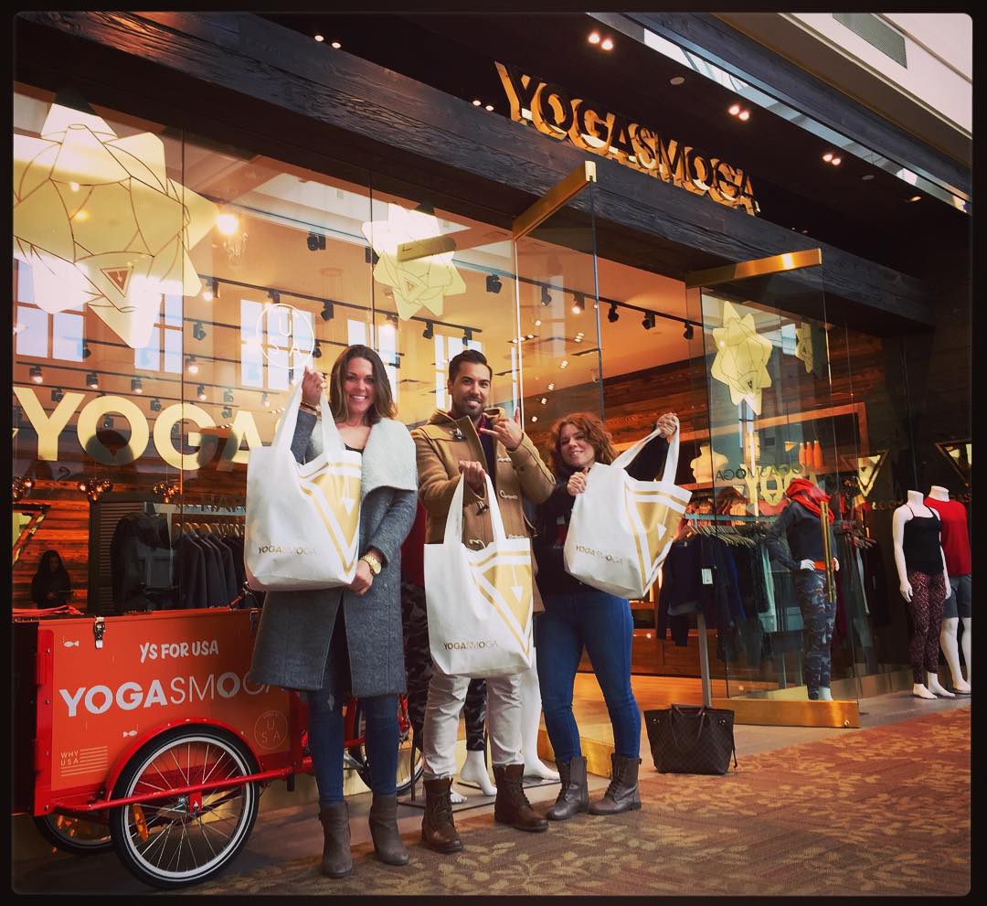 Three shoppers holding up their YS bags in front of one of the retail stores in a mall.