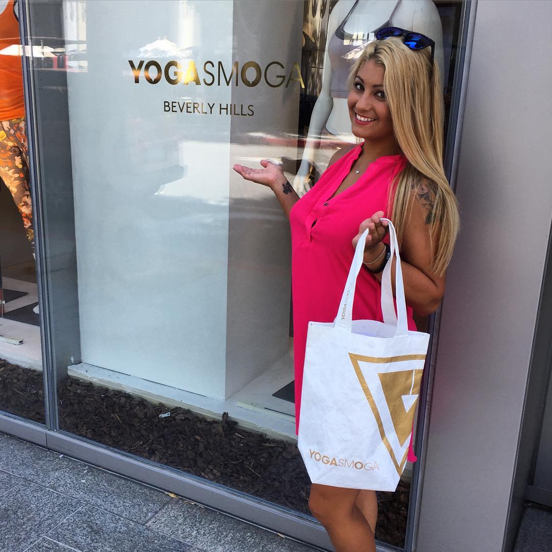 Woman with a pink shirt and YS bag in one hand while gesturing to the YOGASMOGA Beverly Hills gold vinyl sign on the store window.
