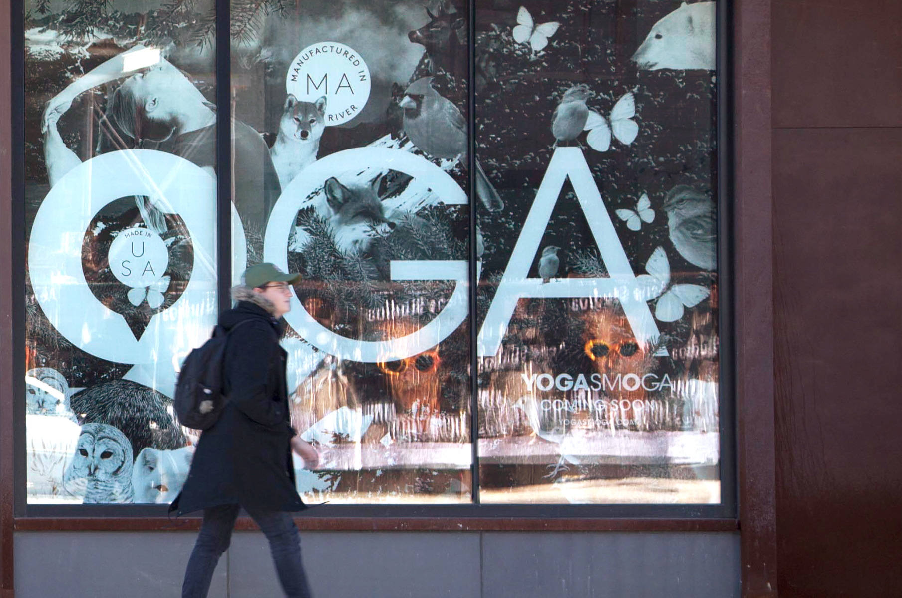 Portion of vinyl collage with giant letters O-G-A surrounded by animals, model in a yoga pose, with a passerby walking in front of the window.