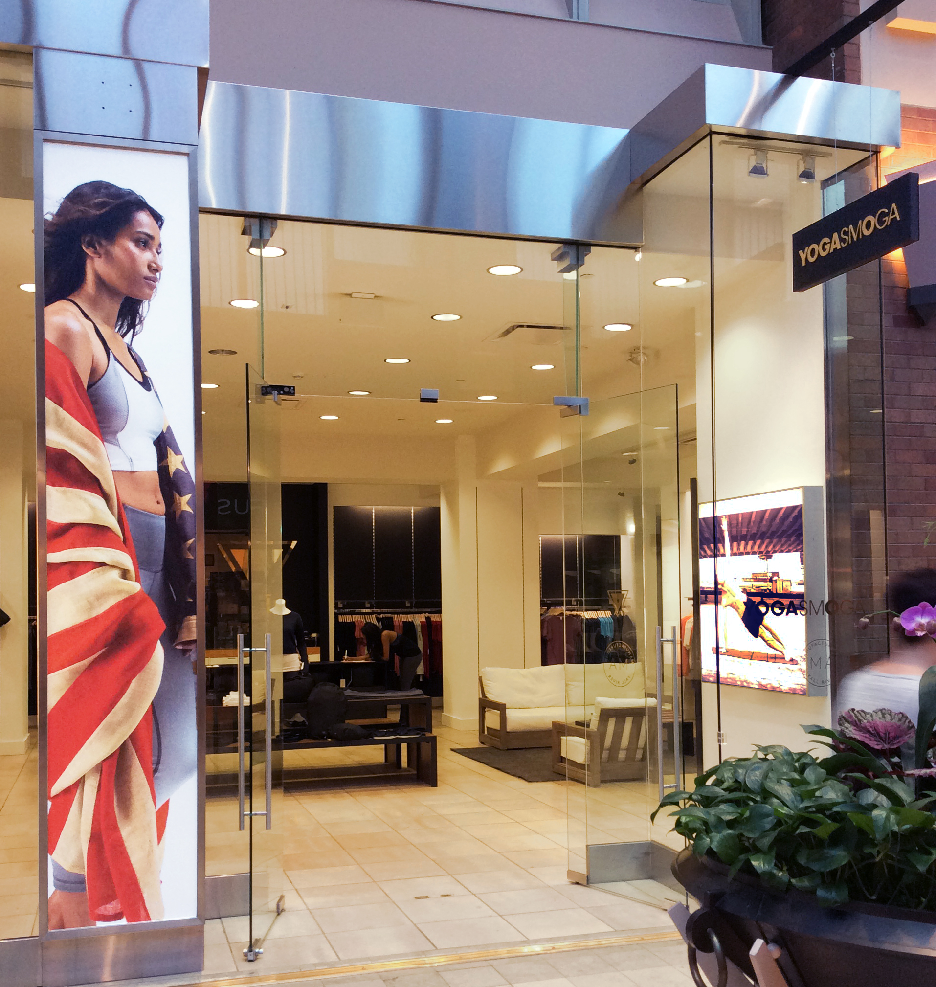 Entrance to the YOGASMOGA Boston store in a mall featuring a tall lightbox showing a model wearing YOGASMOGA yoga clothes draped in an American flag.