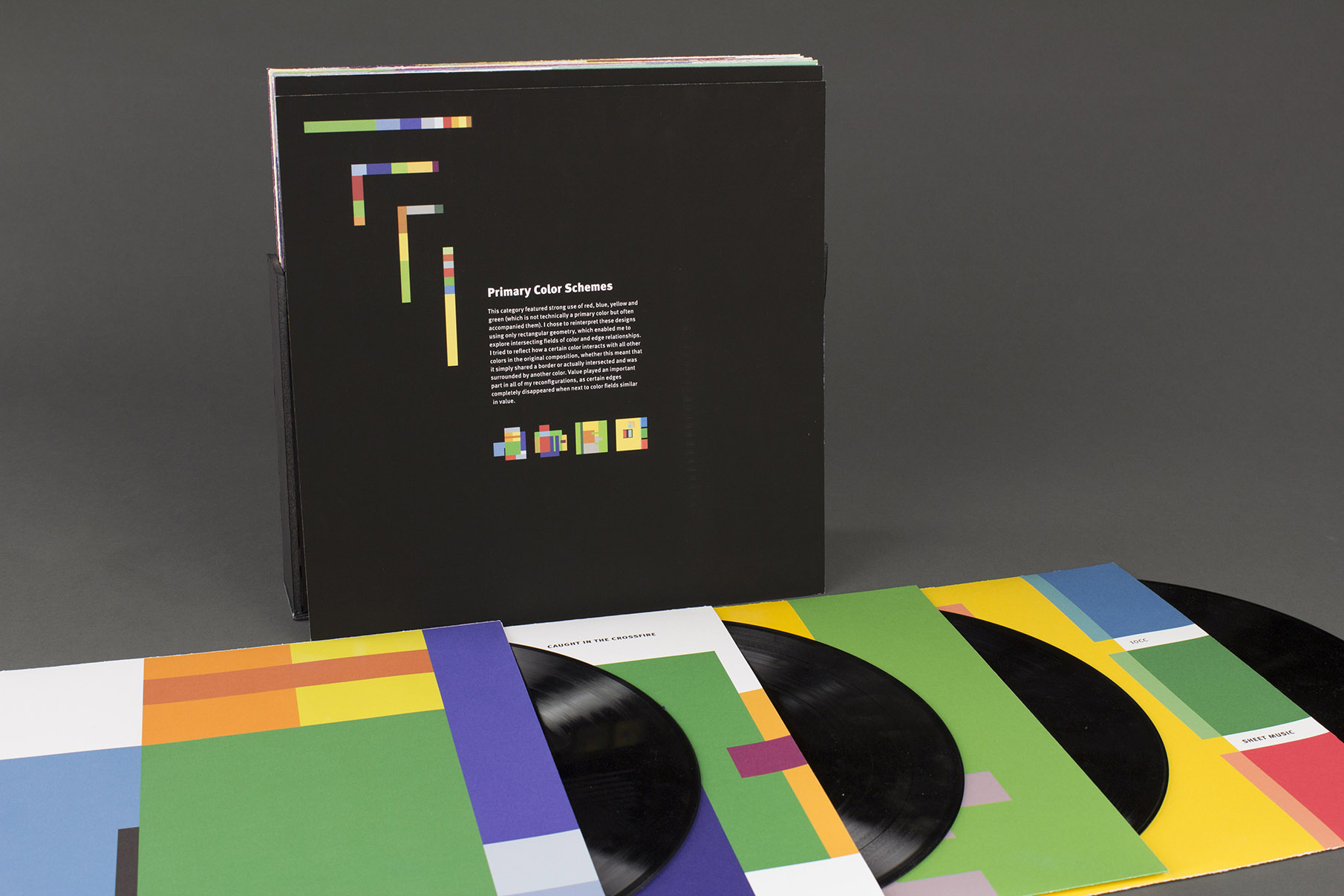 Black album-sized square information card placed upright with the title "Primary Color Schemes" on it. Four vinyl album covers with large color shapes in primary color schemes, and corresponding records arranged in front on the table